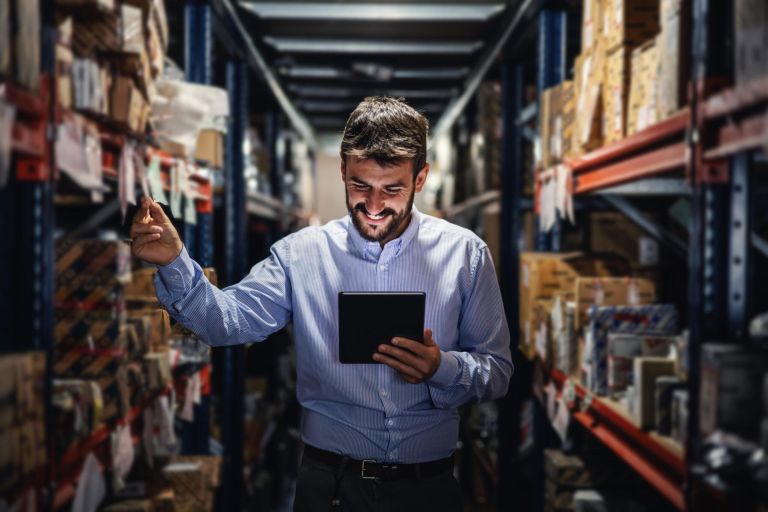 Smiling bearded supervisor standing in warehouse full of boxes and checking on goods ready for deliver.