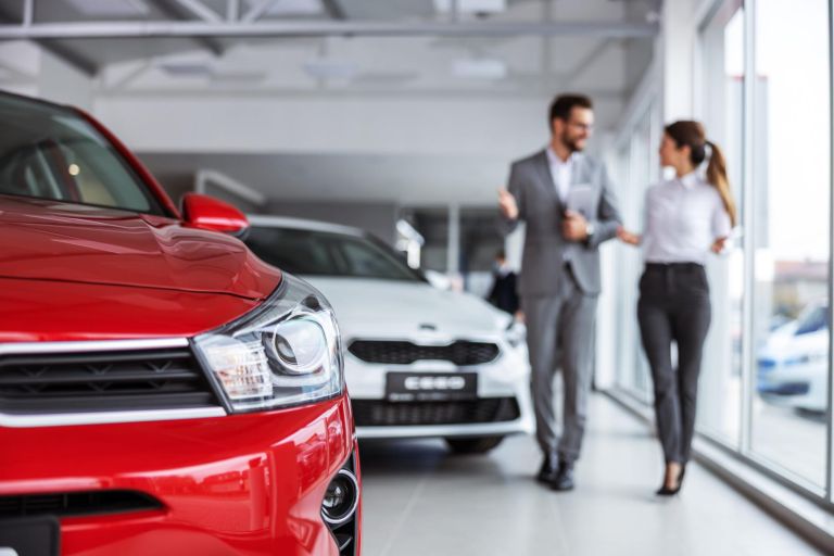 Male car seller in suit walking around car salon with woman who wants to buy a car and talking about specifications of cars.