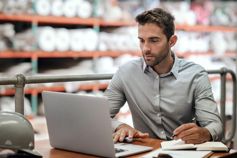 Manager checking online orders and inventory on a laptop while sitting at a desk in a carpet warehouse with shelves of stock in the background