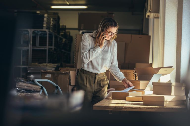 Woman online seller confirming orders from customer on the phone. E-commerce business owner looking at the papers and talking on phone in store warehouse.
